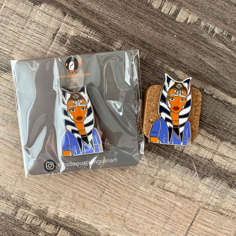 We Love a Little Jumpsuit Moment Ahsoka enamel pin on its backing card next to a pin on cork